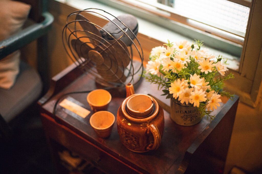 Blooming white and yellow daisy flowers near brown teapot beside desk fan on table